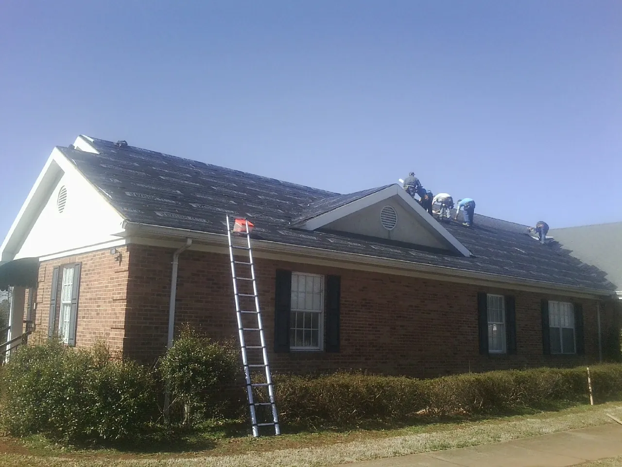 Crew tearing shingles off a brick ranch roof with a ladder set — Emergency Roof Repair work by our crew in Hobgood