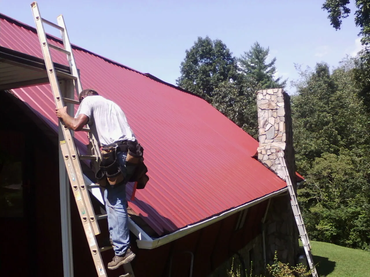 Roofer on a ladder beside a red metal roof and stone chimney — Soffit & Fascia Repair by Roofing Brothers in Hobgood